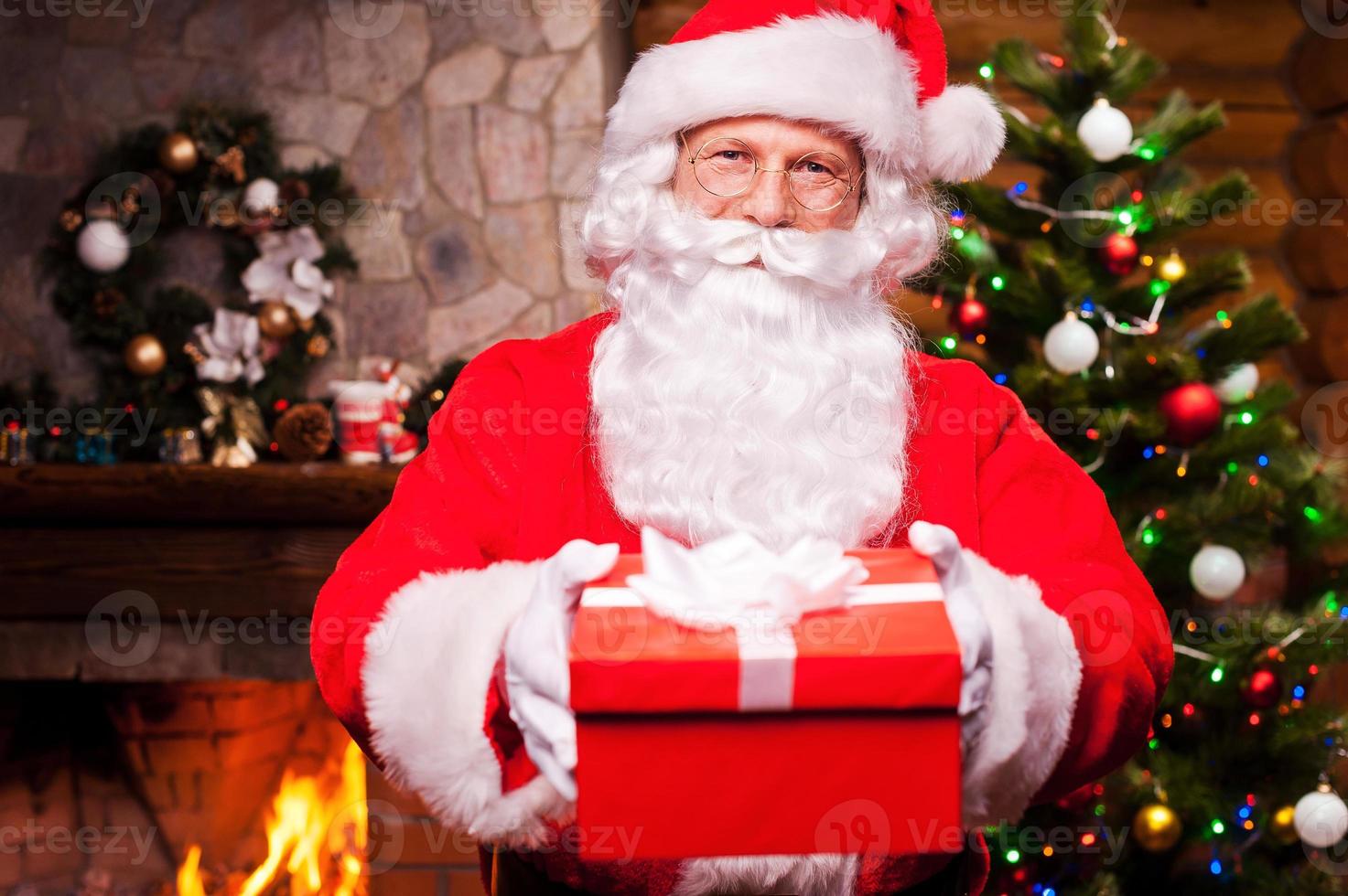 frohe weihnachten für sie fröhlicher weihnachtsmann, der eine geschenkbox ausdehnt und mit kamin und weihnachtsbaum im hintergrund lächelt foto