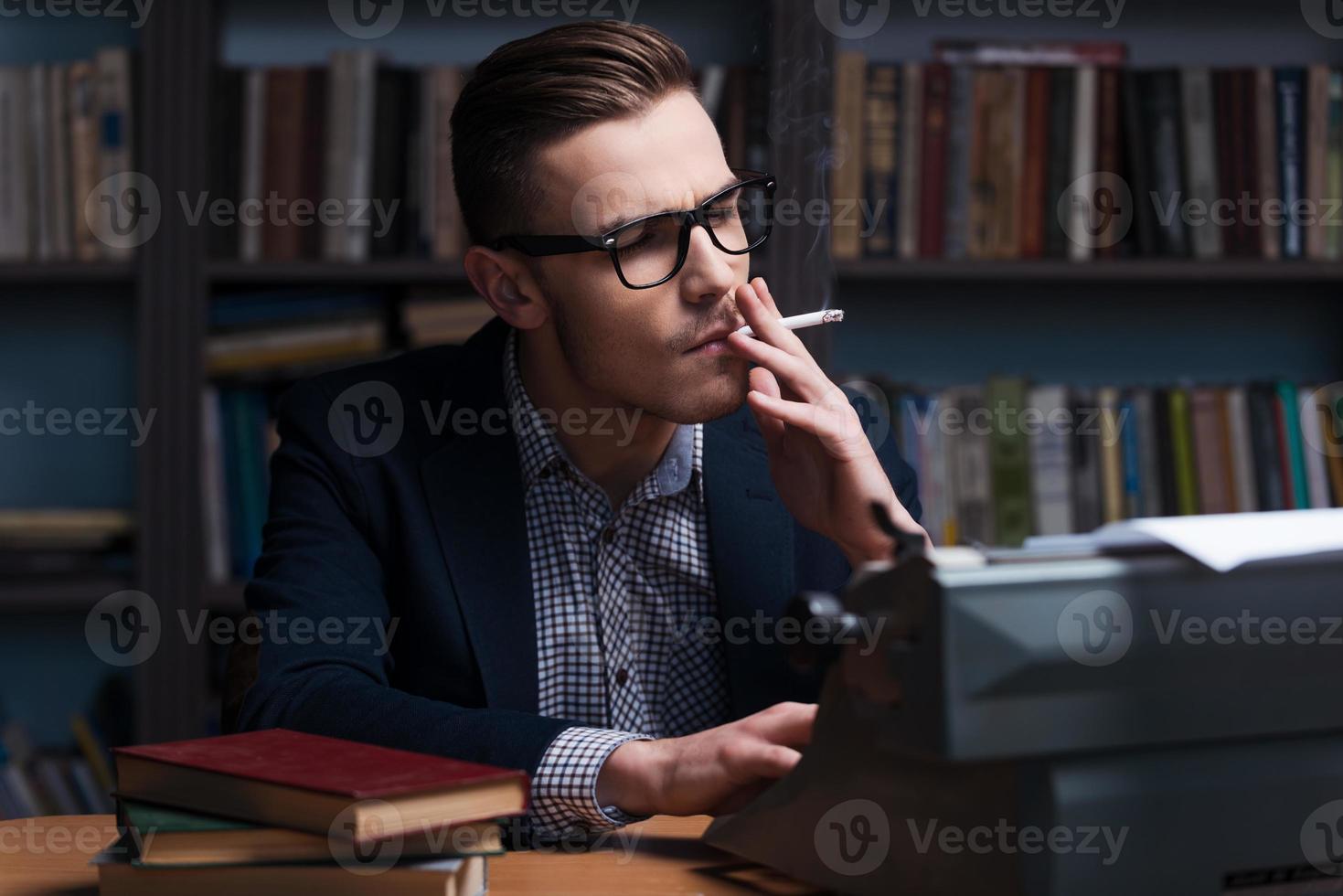 Auf der Suche nach Inspiration. Selbstbewusster junger Autor, der an der Schreibmaschine arbeitet und eine Zigarette raucht, während er an seinem Arbeitsplatz mit einem Bücherregal im Hintergrund sitzt foto