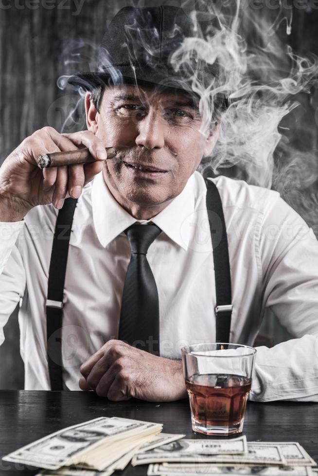 Mafia schläft nicht. ernsthafter älterer Mann in Hemd und Hosenträgern, der am Tisch sitzt und Zigarre raucht, während viel Geld neben ihm liegt foto