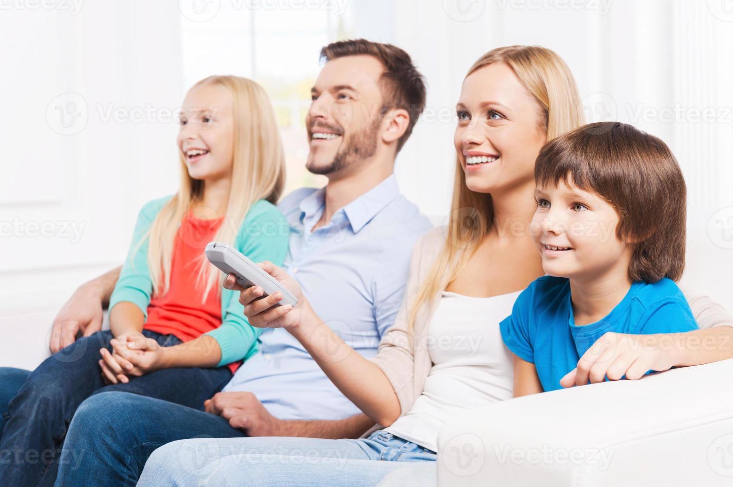 ihre Lieblingssendung ansehen. Seitenansicht einer glücklichen vierköpfigen Familie, die sich aneinander bindet und lächelt, während sie zu Hause zusammen fernsieht foto