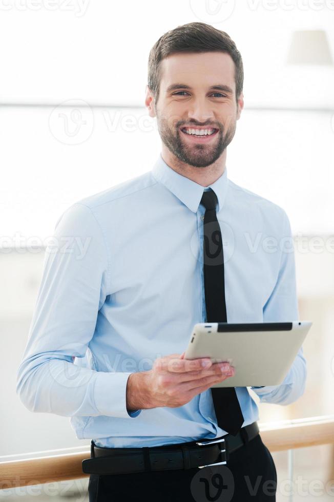 Geschäftsmann mit Notizblock. Fröhlicher junger Geschäftsmann, der ein digitales Tablet hält und lächelt, während er drinnen steht foto