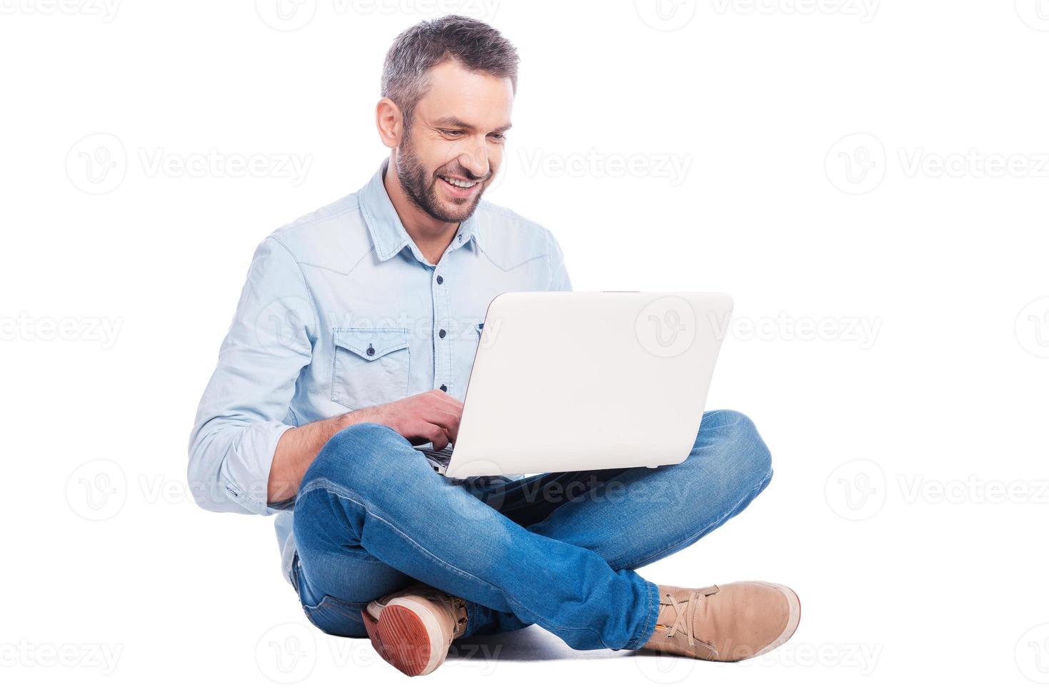 kostenloses WLAN genießen. hübscher junger Mann in Freizeitkleidung, der auf dem Boden sitzt und am Laptop arbeitet, während er isoliert auf weißem Hintergrund ist foto