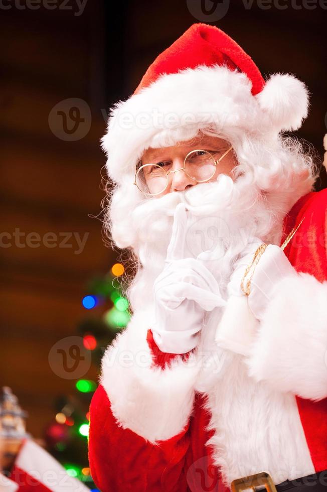 shhh traditioneller weihnachtsmann gestikuliert schweigezeichen, während er sack mit geschenken und mit weihnachtsbaum im hintergrund trägt foto