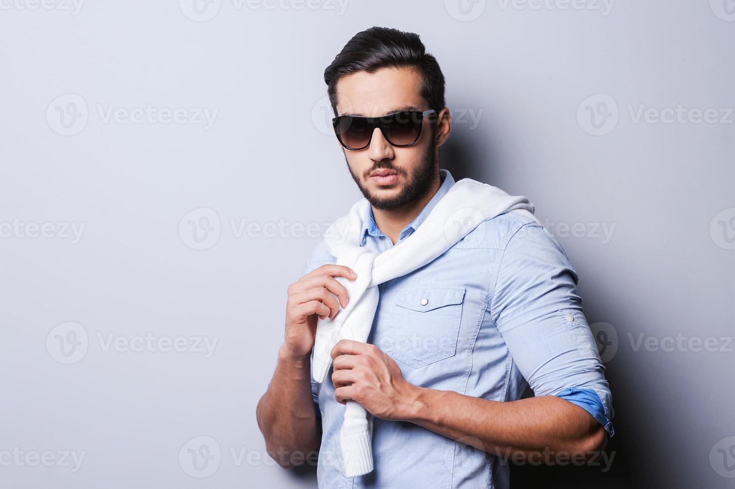 lässig cool. hübscher junger Mann im blauen Hemd, der Pullover auf ihren Schultern bindet, während er vor grauem Hintergrund steht foto