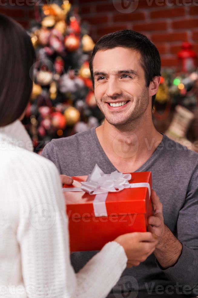 Was für eine überraschende Rückansicht des jungen Mannes, der seiner Freundin eine rote Geschenkbox mit Weihnachtsbaum im Hintergrund gibt foto
