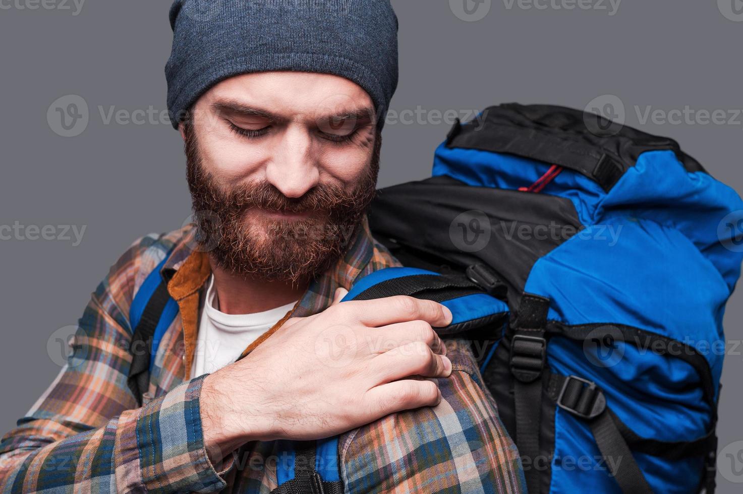 Rucksack zu schwer. Frustrierter junger bärtiger Mann, der seinen Rucksack anpasst, während er vor grauem Hintergrund steht foto