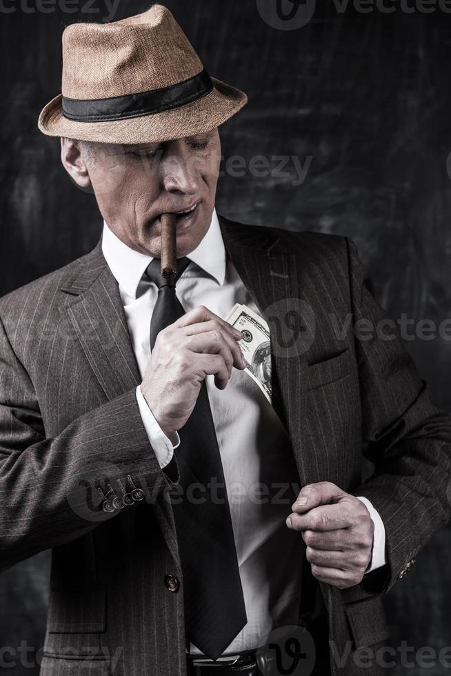 Geld und Macht. Ernsthafter älterer Mann mit Hut und Hosenträgern, der Zigarre raucht und Geld in der Tasche versteckt, während er vor dunklem Hintergrund steht foto