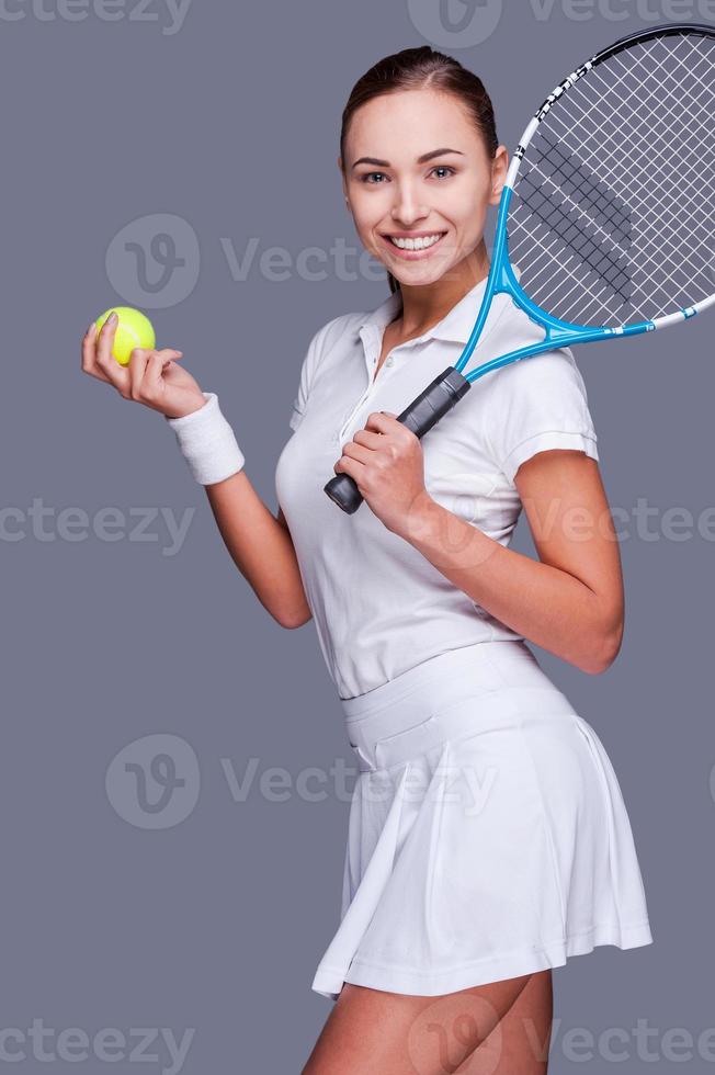 bringt die Schönheit ins Spiel. Schöne junge Frauen in Sportkleidung, die Tennisschläger auf der Schulter halten und im Stehen vor grauem Hintergrund lächeln foto