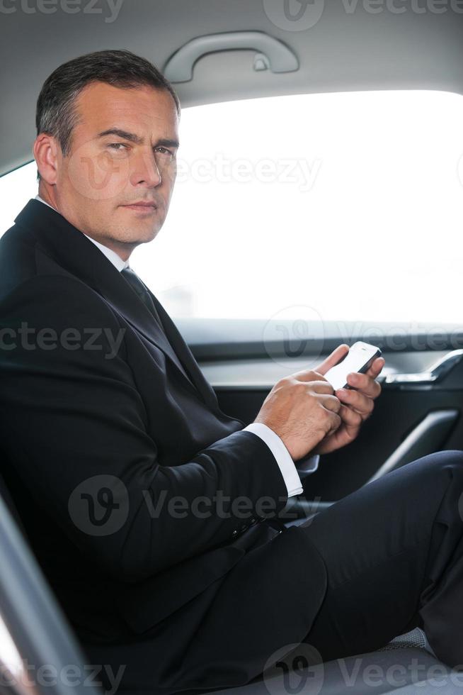 eine geschäftliche Nachricht eingeben. Seitenansicht eines selbstbewussten, reifen Geschäftsmanns, der eine Nachricht auf seinem Smartphone tippt und in die Kamera schaut, während er auf dem Rücksitz eines Autos sitzt foto