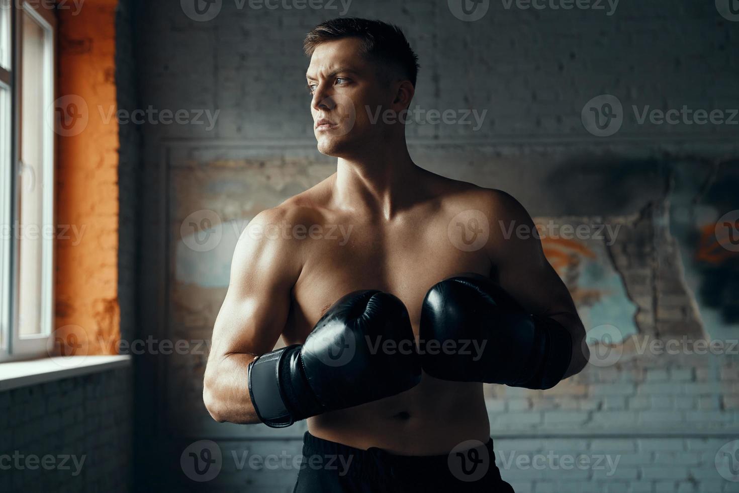 Selbstbewusster junger Mann in Boxhandschuhen, der wegschaut, während er im Fitnessstudio steht foto