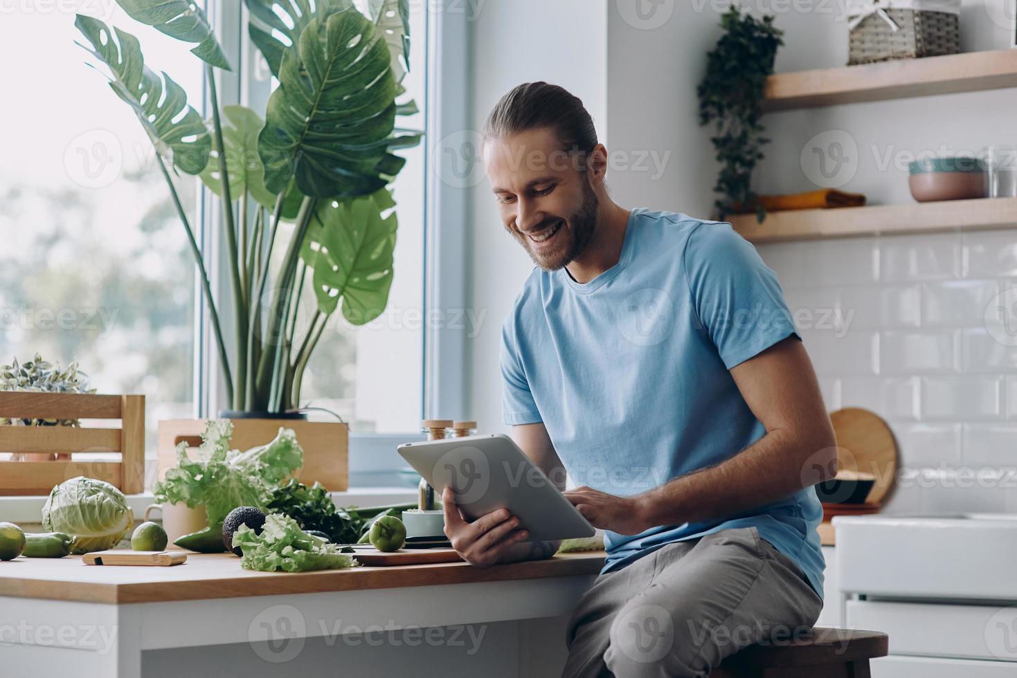 Fröhlicher junger Mann, der ein digitales Tablet hält, während er in der heimischen Küche Essen zubereitet foto