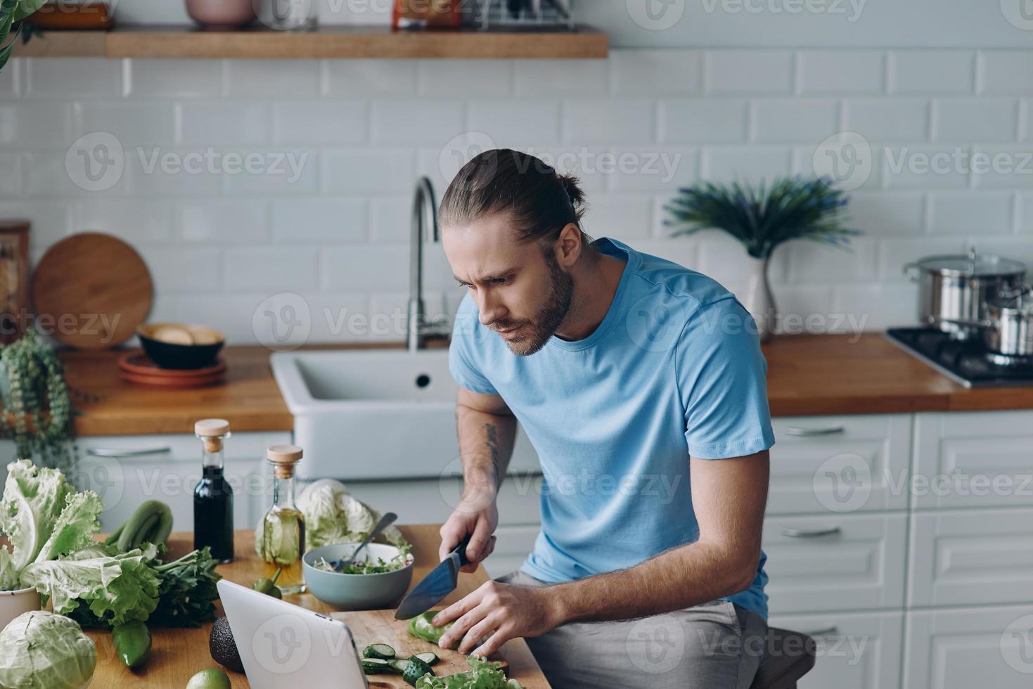 gutaussehender junger Mann, der sich ein digitales Tablet ansieht, während er in der heimischen Küche Essen zubereitet foto