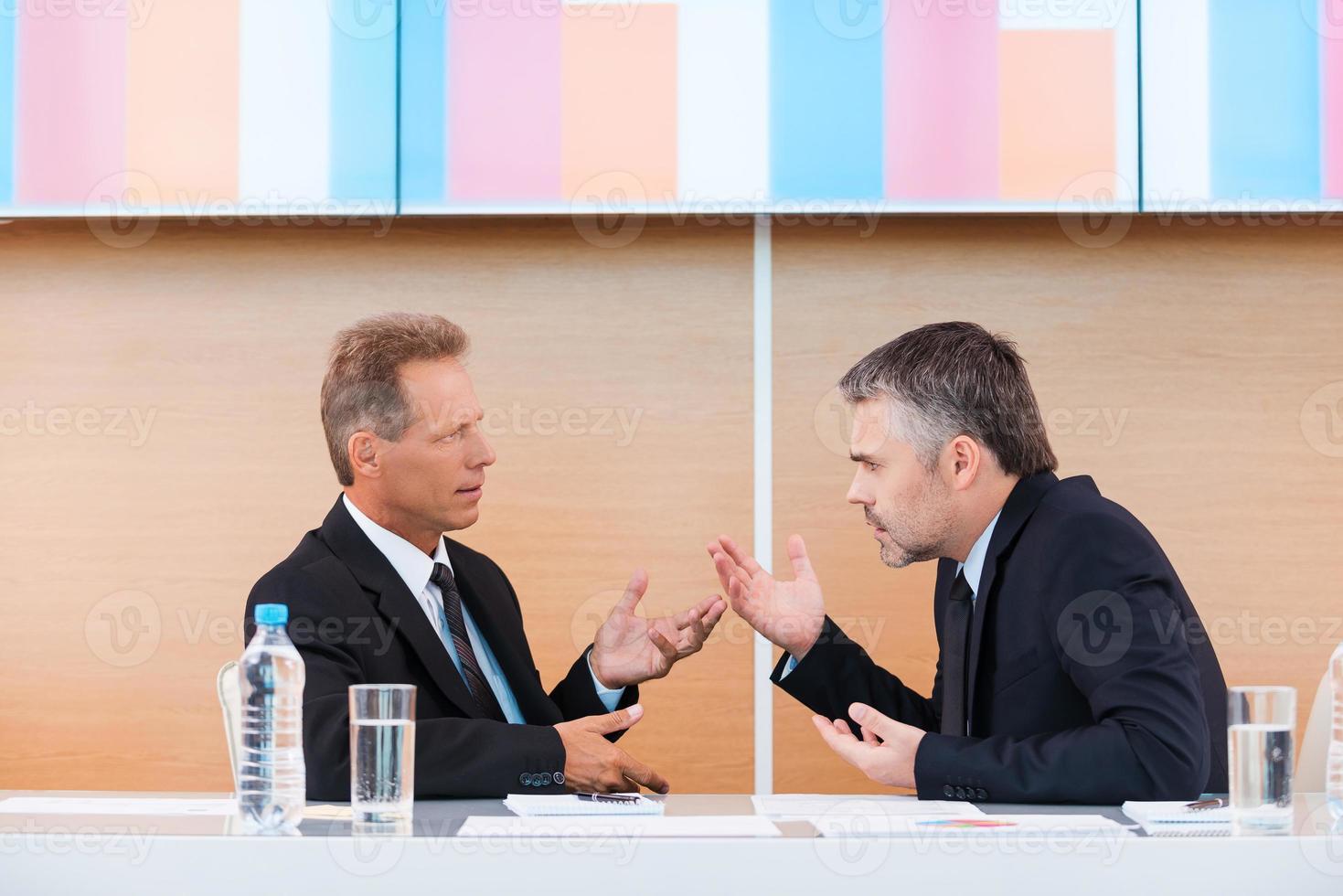 heiße Diskussion. Zwei wütende Geschäftsleute in Abendgarderobe streiten und gestikulieren, während sie mit einem großen Monitor zusammen am Tisch sitzen foto