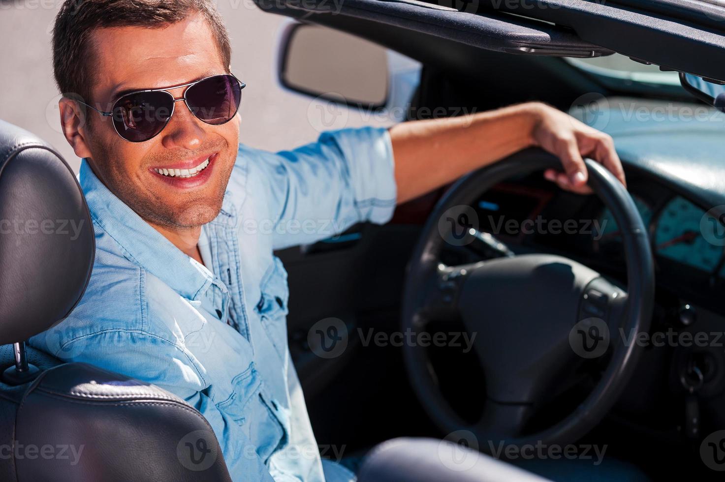 bereit zu gehen. hübscher junger Mann mit Sonnenbrille, der auf dem Vordersitz seines Cabrios sitzt und lächelt foto