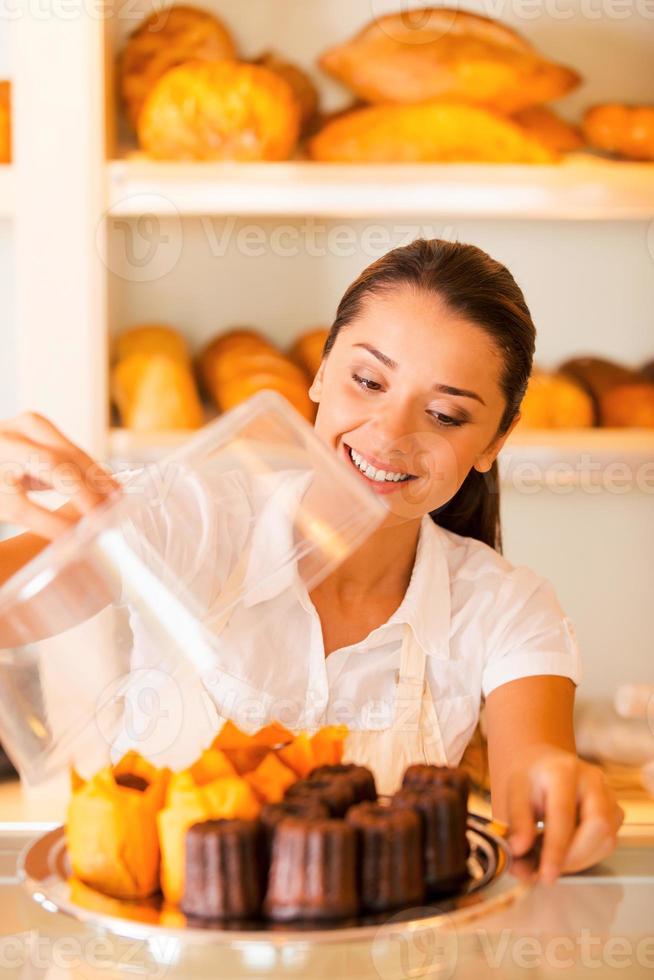 handgemachte Kekse zu verkaufen. Schöne junge Frau in Schürze, die Teller mit frischen Keksen trägt und lächelt, während sie in der Bäckerei steht foto
