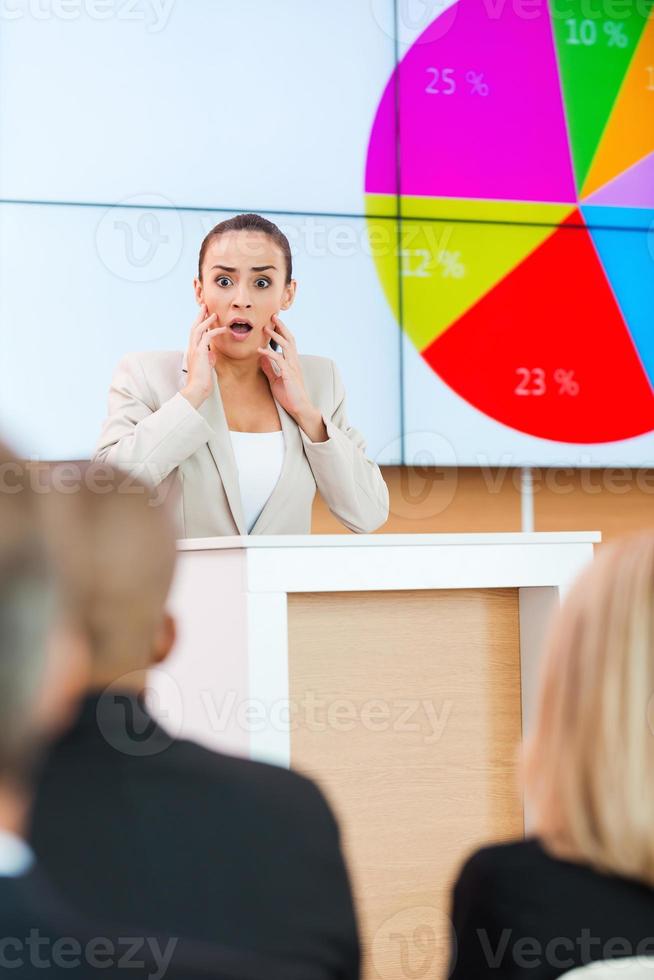 Angst vor öffentlichem Redner. Schockierte junge Frau in Abendkleidung, die ihr Gesicht mit den Händen berührt, während sie auf der Tribüne im Konferenzsaal mit Menschen im Vordergrund steht foto