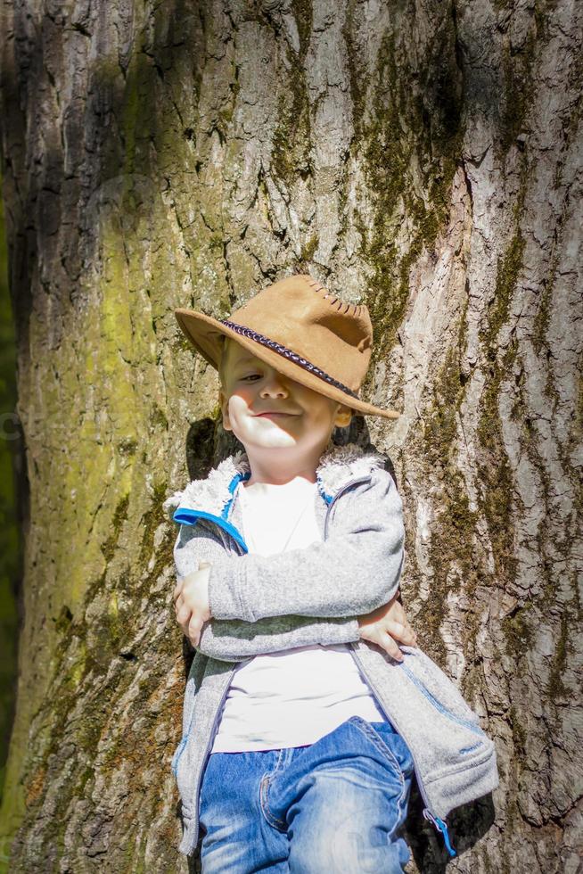 Süßer Junge, der in einem Cowboyhut im Wald bei einem Baum posiert. die Sonnenstrahlen umhüllen den Raum. Interaktionsgeschichte für das Buch. Platz zum Kopieren foto