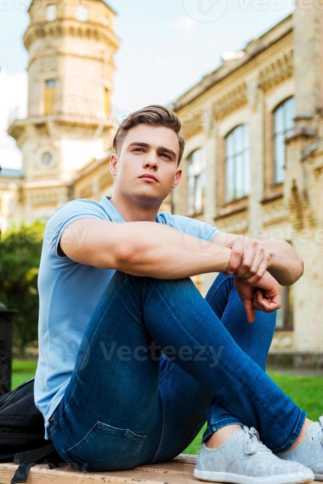 an die Zukunft denken. nachdenklicher männlicher Student, der auf der Bank sitzt und mit dem Universitätsgebäude im Hintergrund wegschaut foto