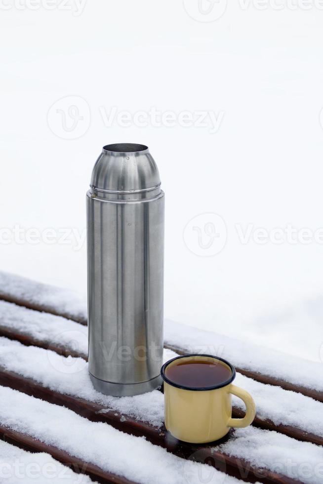 Thermoskanne und Metallbecher mit heißem Tee auf einer schneebedeckten Bank in einem Winterpark. foto