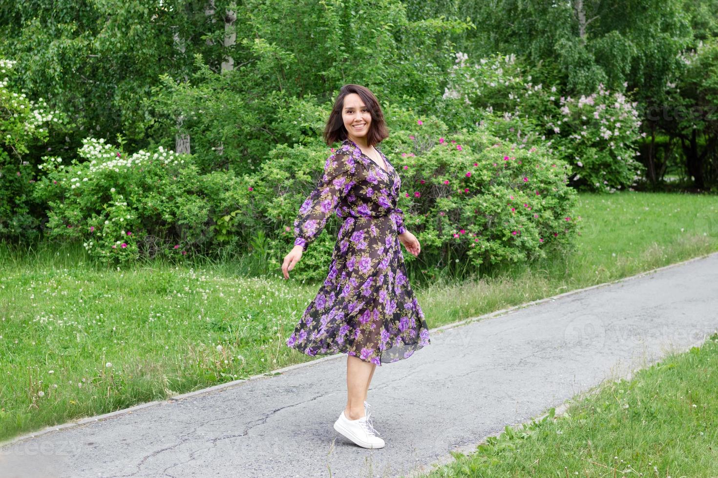 Schöne junge Frau in schwarz-violettem Kleid geht in einem Garten mit blühenden Fliederbüschen spazieren. foto