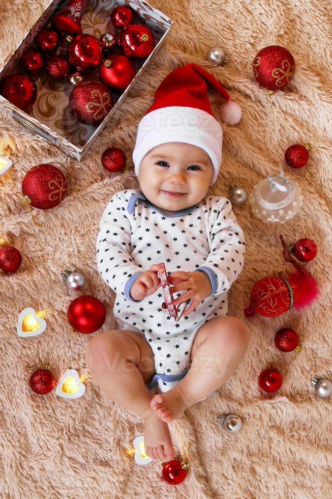 Schönes lächelndes kleines Mädchen im roten Hut des Weihnachtsmanns spielt mit Holzspielzeug auf einem beigefarbenen Plaid mit roten und weißen Weihnachtsdekorationen und Weihnachtslichtern, Draufsicht. foto
