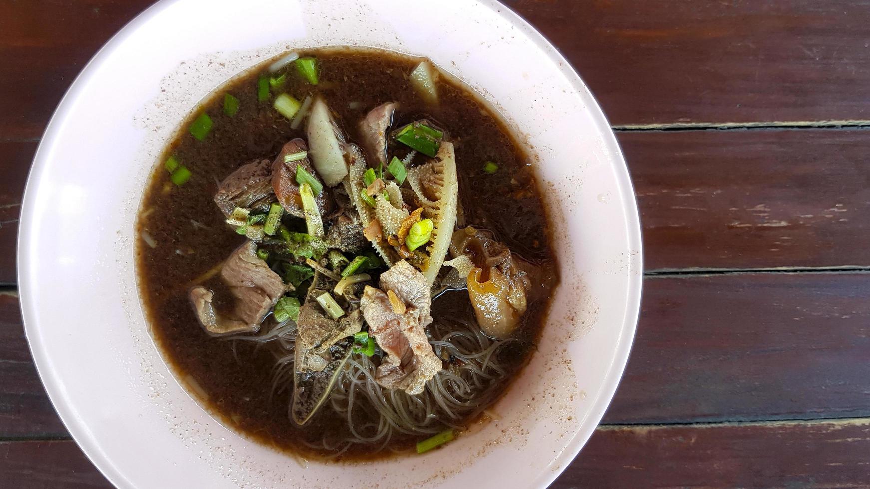 Draufsicht auf thailändische Reisnudeln mit Rindfleisch, abgestandenem Fleisch, Leber, Kuhlappen, Innereien und auf braunem Holzhintergrund oder Tisch und Kopienraum rechts bei lokalem Straßenessen. flache lage asiatischer speisen auf rosa schüssel. foto