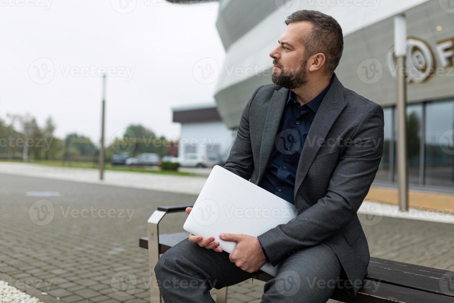 Porträt eines erfolgreichen reifen Geschäftsmannes mit Laptop, der neben einem Bürogebäude sitzt foto