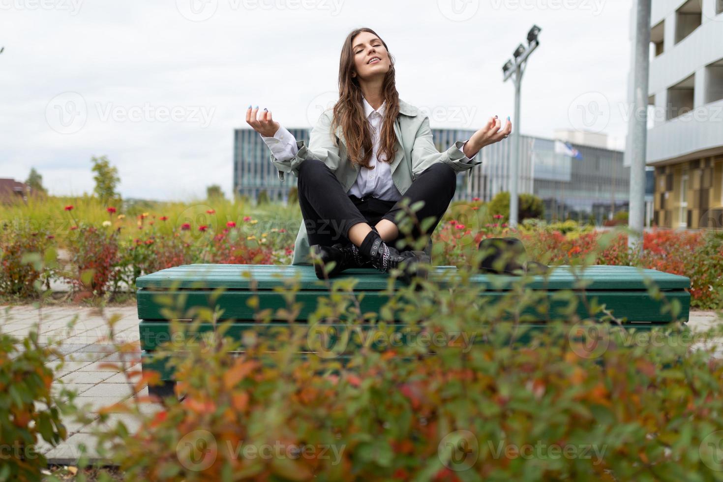 Eine junge Frau sitzt auf einer Bank im Park in einer Yoga-Pose mit gespreizten Armen und geschlossenen Augen foto
