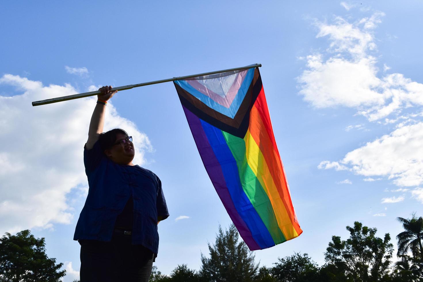 asiatischer junger teenager im schwarzen hemd hält lgbtq-flagge in den händen und winkt, weicher und selektiver fokus, verschwommener wolkiger und blauer hintergrund, konzept für lgbtq-geschlechterfeiern im stolzmonat. foto