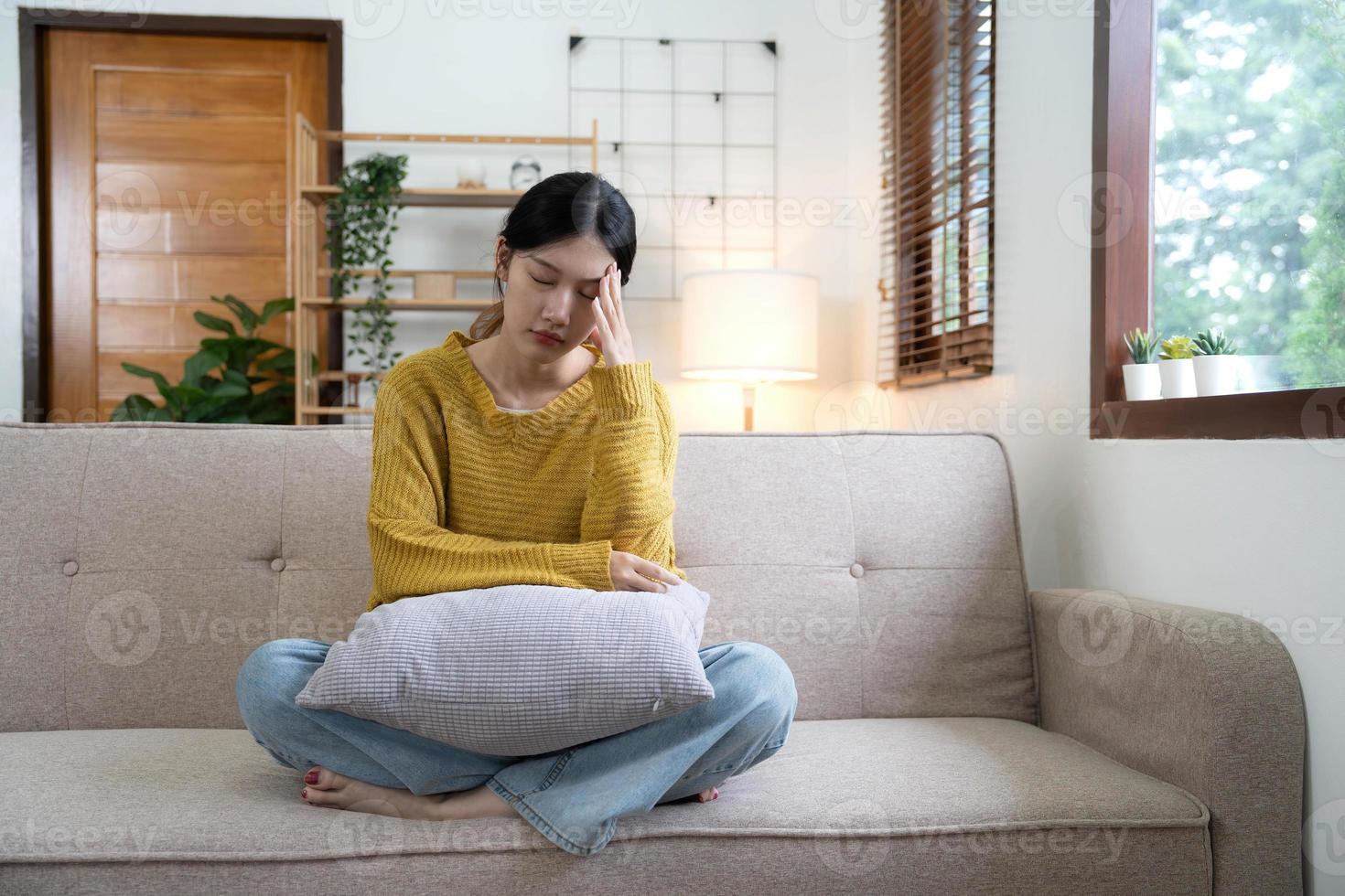 Hand auf Schläfen junger unglücklicher Traurigkeit asiatisches Mädchen, das auf dem Sofa sitzt. sie fühlt sich wegen ihrer krankheit nicht sehr gut und hat kopfschmerzen. von vorne gesehen. stress- und krankheitskonzept. foto