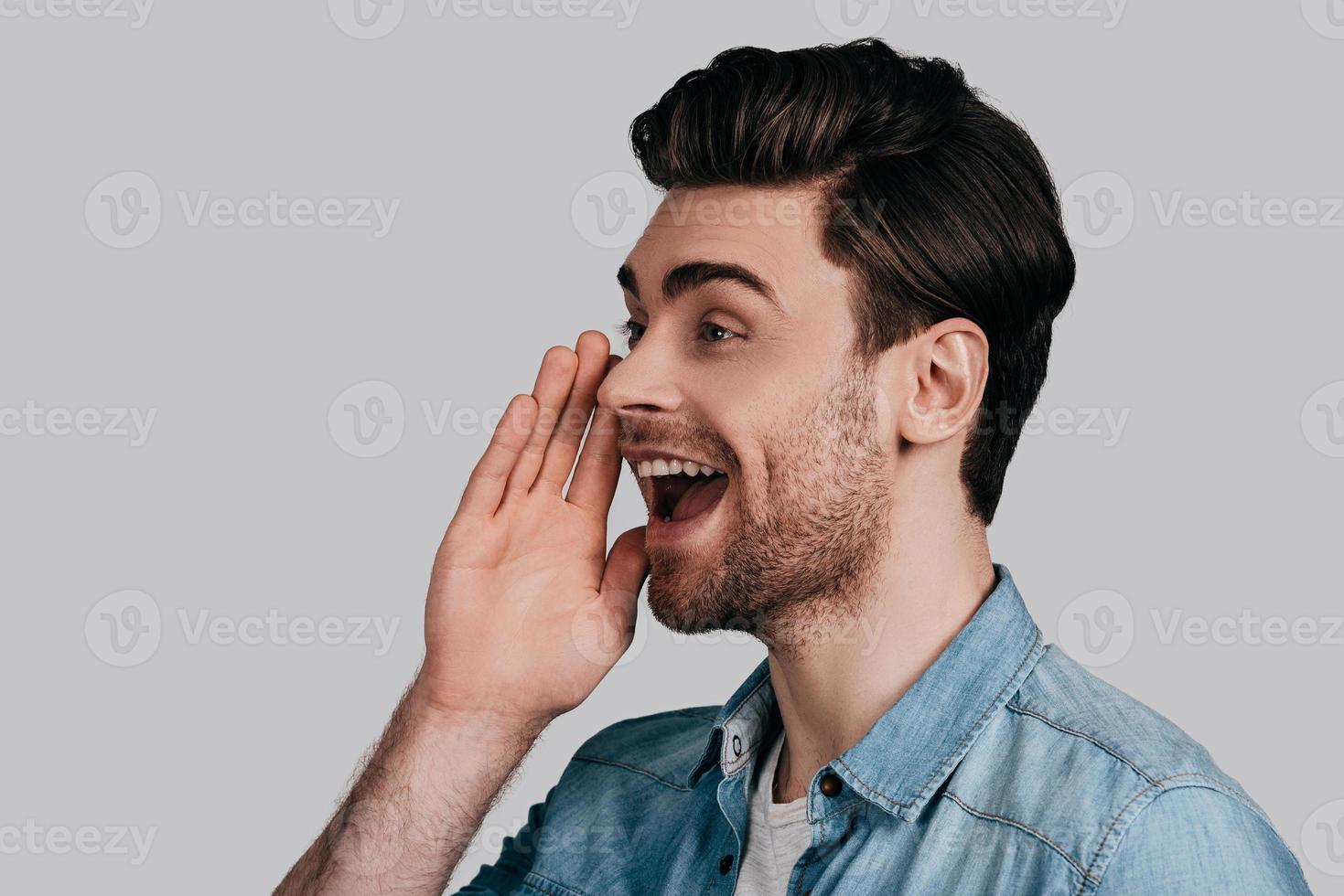 gute Nachrichten teilen. Hübscher junger Mann im Blue Jeans-Hemd, der schreit und die Hand über der Maus hält, während er vor grauem Hintergrund steht foto