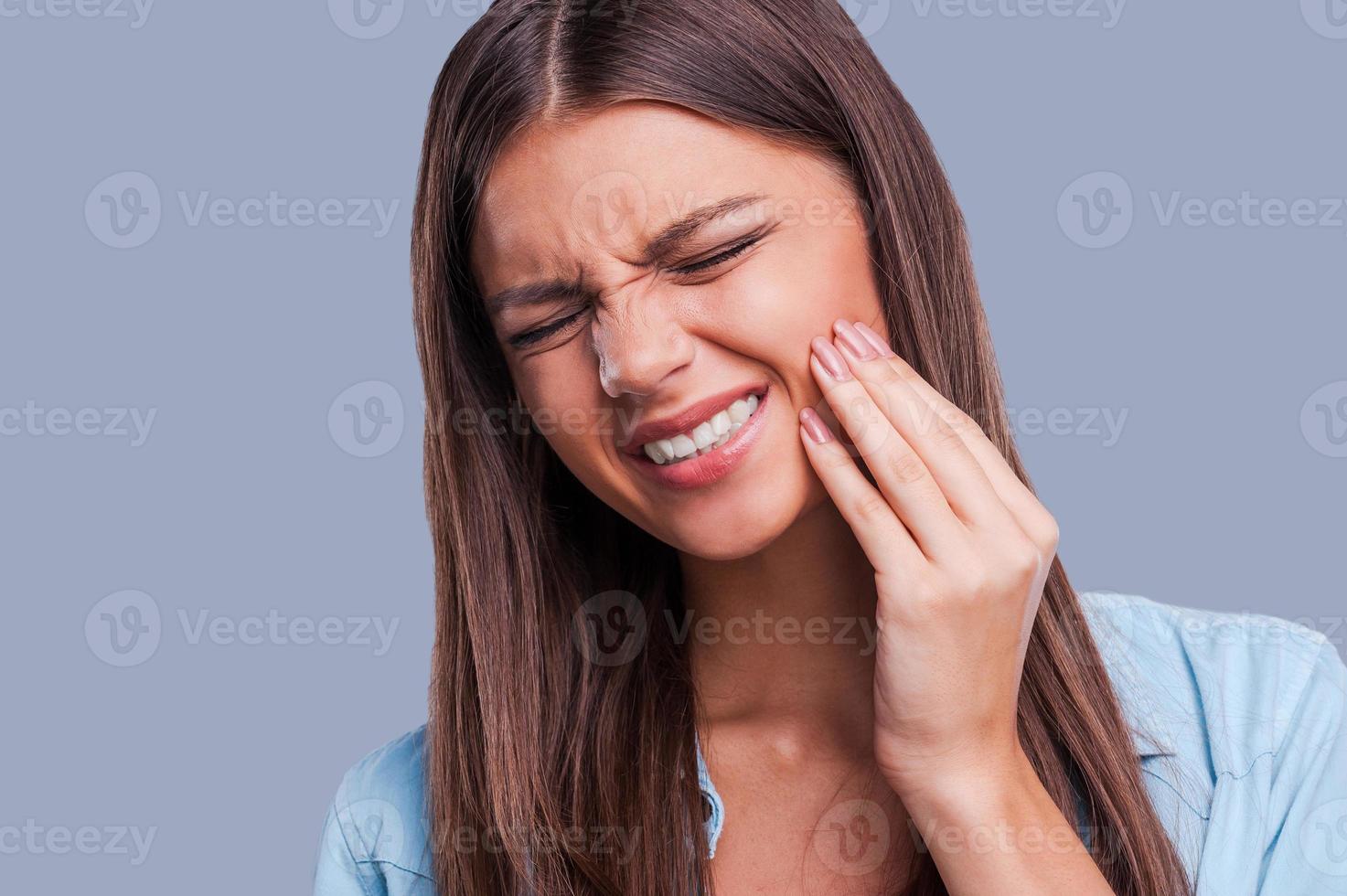 unter Zahnschmerzen leiden. schöne junge Frau, die unter Zahnschmerzen leidet, während sie vor grauem Hintergrund steht foto