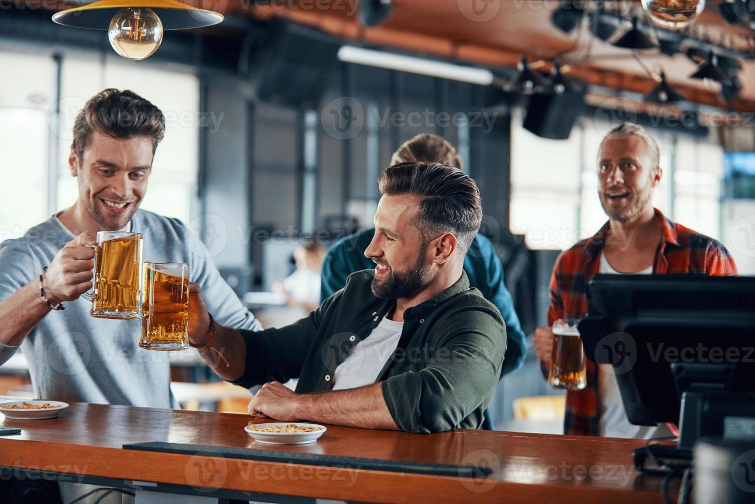 Jubelnde junge Männer in Freizeitkleidung, die Sportspiele verfolgen und Bier genießen, während sie in der Kneipe sitzen foto