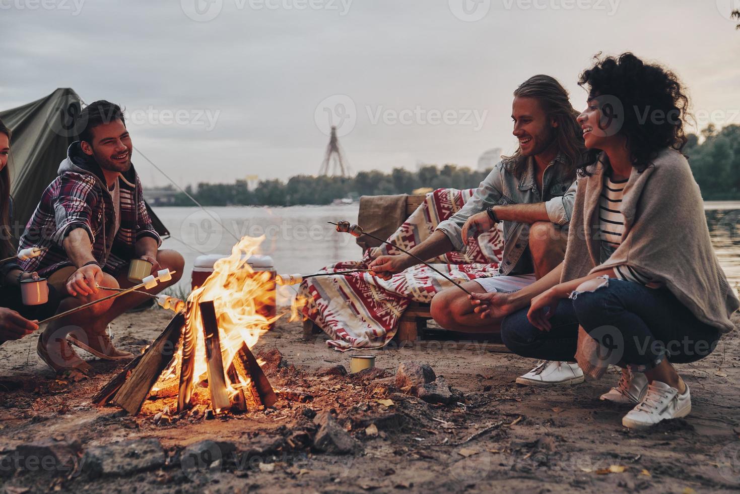 keine Sorgen. Gruppe junger Leute in Freizeitkleidung, die Marshmallows über einem Lagerfeuer rösten, während sie sich in der Nähe des Sees ausruhen foto