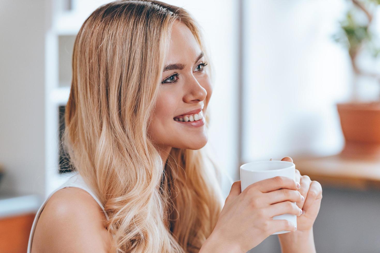 lächelnde Schönheit. Nahaufnahme der schönen, fröhlichen jungen Frau, die eine Kaffeetasse hält und mit einem Lächeln wegschaut, während sie zu Hause in der Küche sitzt foto