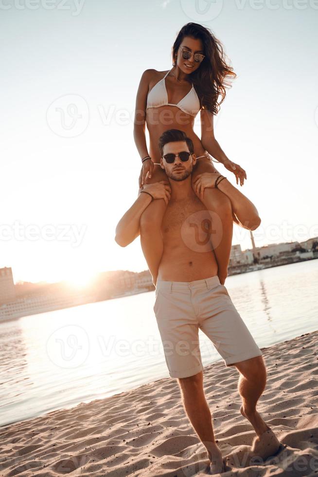 Liebe liegt in der Luft. in voller Länge von einem hübschen jungen Mann, der seine attraktive Freundin auf den Schultern trägt, während er am Strand steht foto