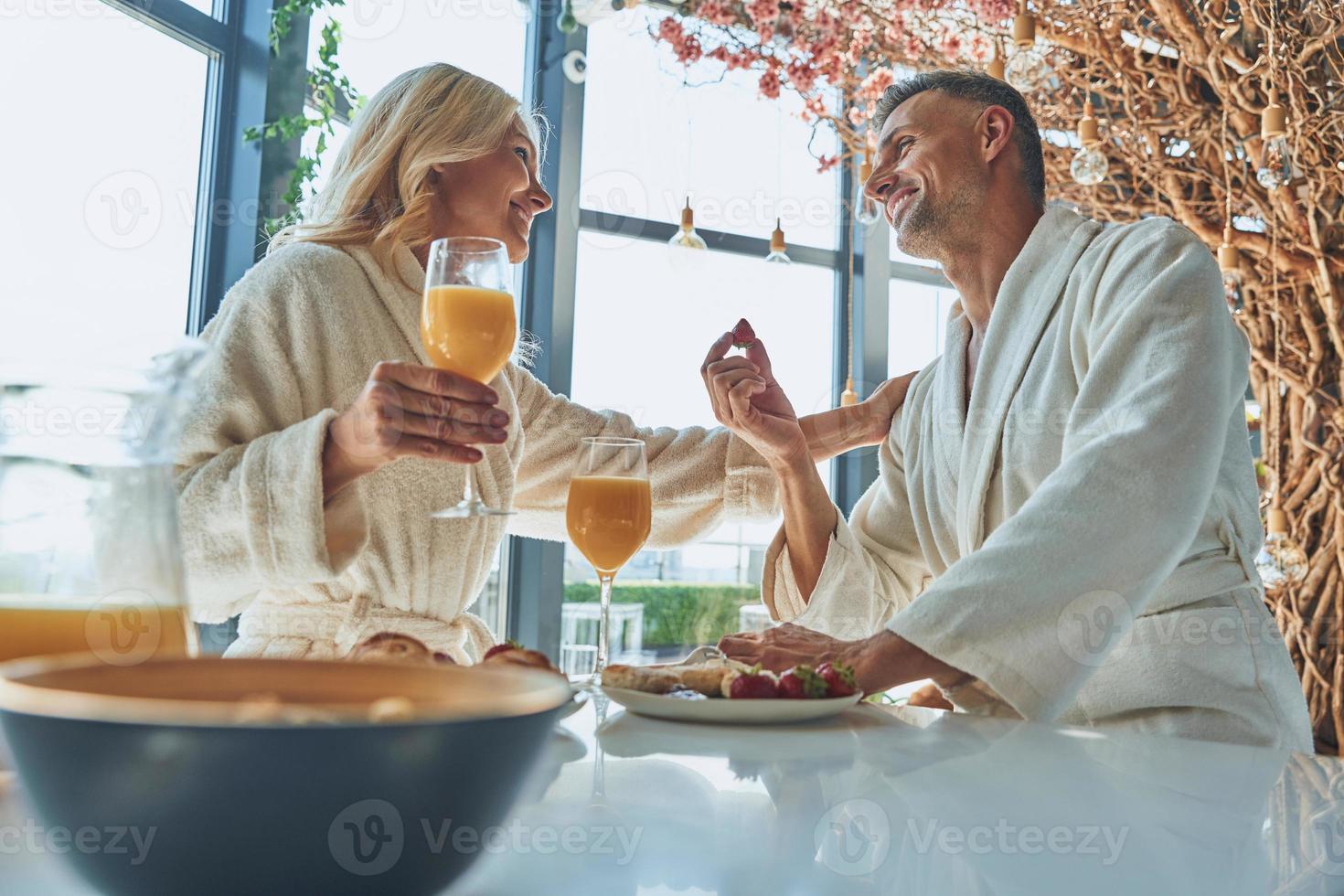 Schönes reifes Paar in Bademänteln, das zusammen frühstückt, während es Zeit in der heimischen Küche verbringt foto