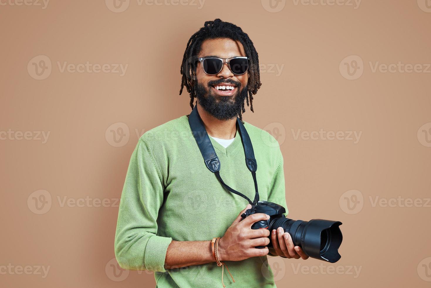 junger afrikanischer Fotograf in Freizeitkleidung, der in die Kamera schaut und lächelt, während er vor braunem Hintergrund steht foto