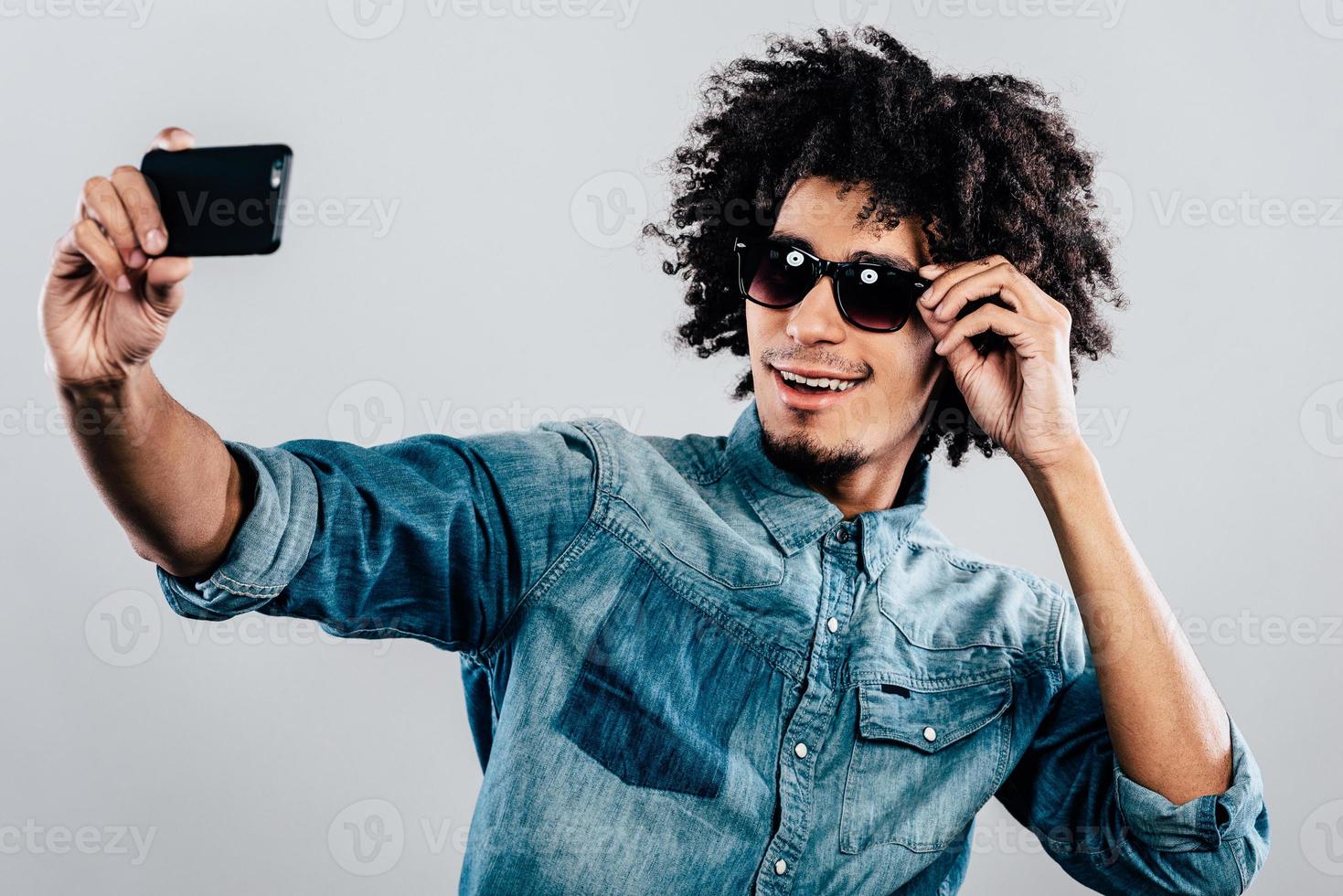 stylisches Selfie. Fröhlicher junger afrikanischer Mann mit Sonnenbrille und Selfie im Stehen vor grauem Hintergrund foto