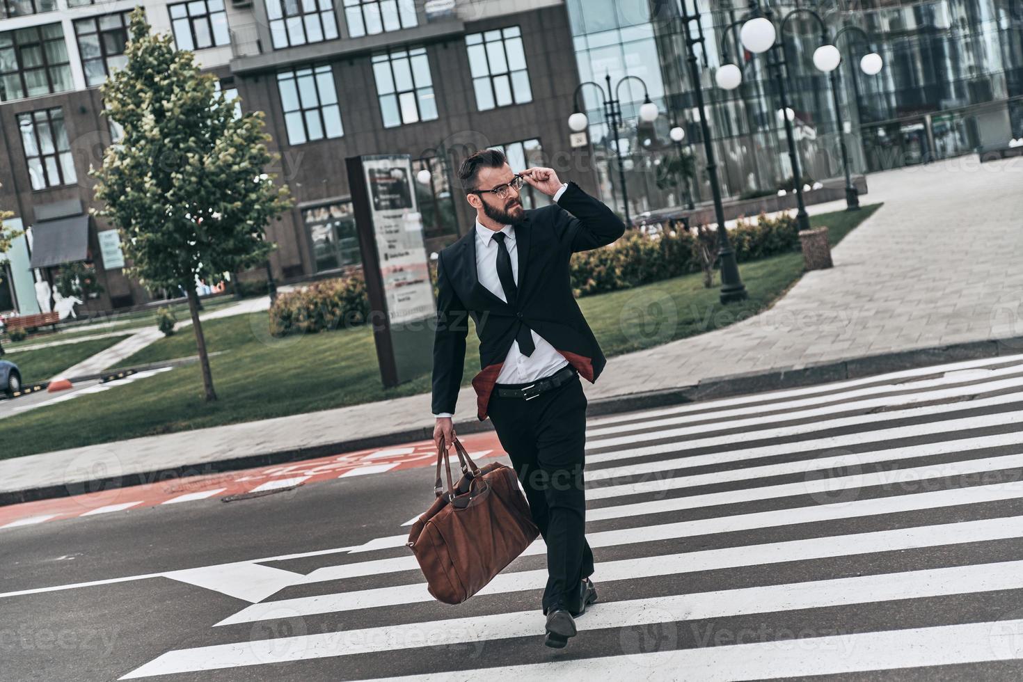 selbstbewusster Geschäftsmann. voller Länge des jungen Mannes im vollen Anzug, der die Straße beim Gehen im Freien überquert foto