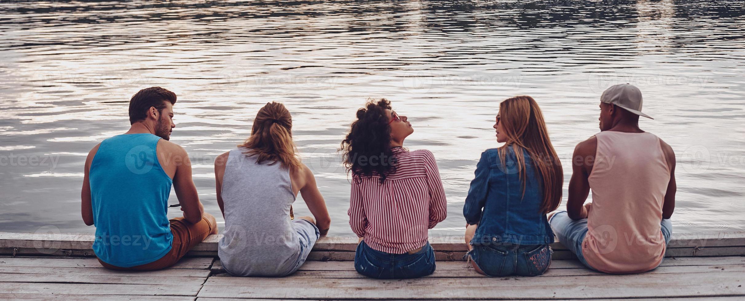 unbeschwerte Zeit mit Freunden. Rückansicht junger Menschen in Freizeitkleidung, die sich unterhalten, während sie auf dem Pier sitzen foto