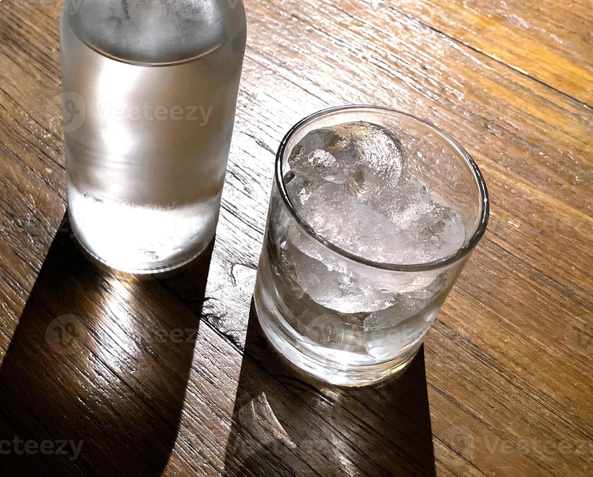 Selektiver Fokus, ein mit Eis gefülltes klares Glas und eine klare Glasflasche hält kaltes Mineralwasser auf einem Holztisch foto