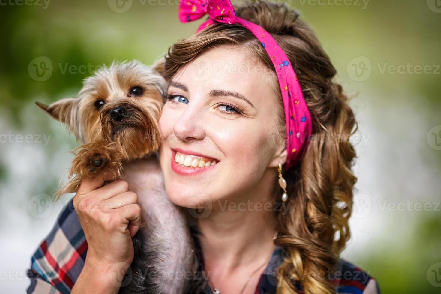 Schönes junges Mädchen in einem karierten Hemd und kurzen Jeansshorts im Pin-up-Stil im Wald mit einem kleinen Hund des Yorkshire-Terriers in ihren Armen foto