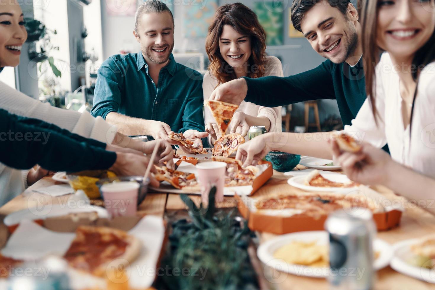 Gruppe junger Leute in Freizeitkleidung, die Pizza pflücken und lächeln, während sie drinnen eine Dinnerparty haben foto