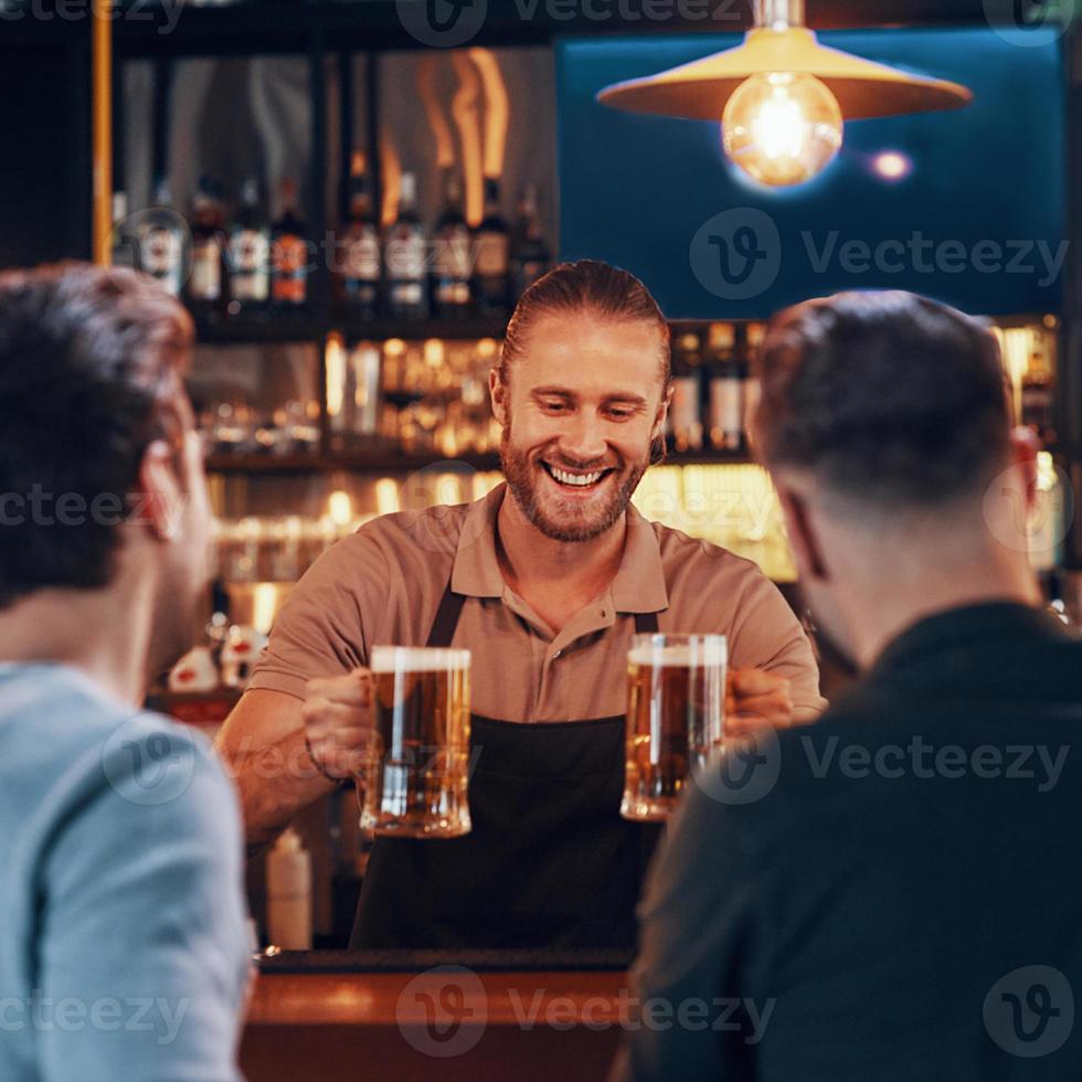 Fröhlicher Barkeeper, der jungen Männern Bier serviert, während er an der Bartheke im Pub steht foto