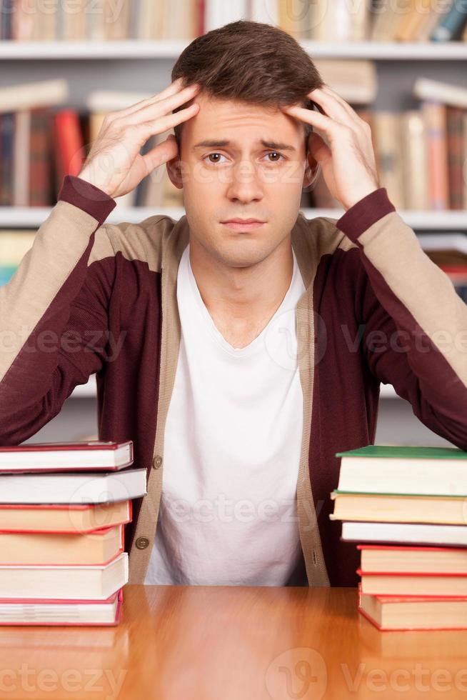 müde vom Lernen. müder junger mann, der seinen kopf in den händen hält und sich an den buchstapel lehnt, während er am bibliotheksschreibtisch sitzt foto