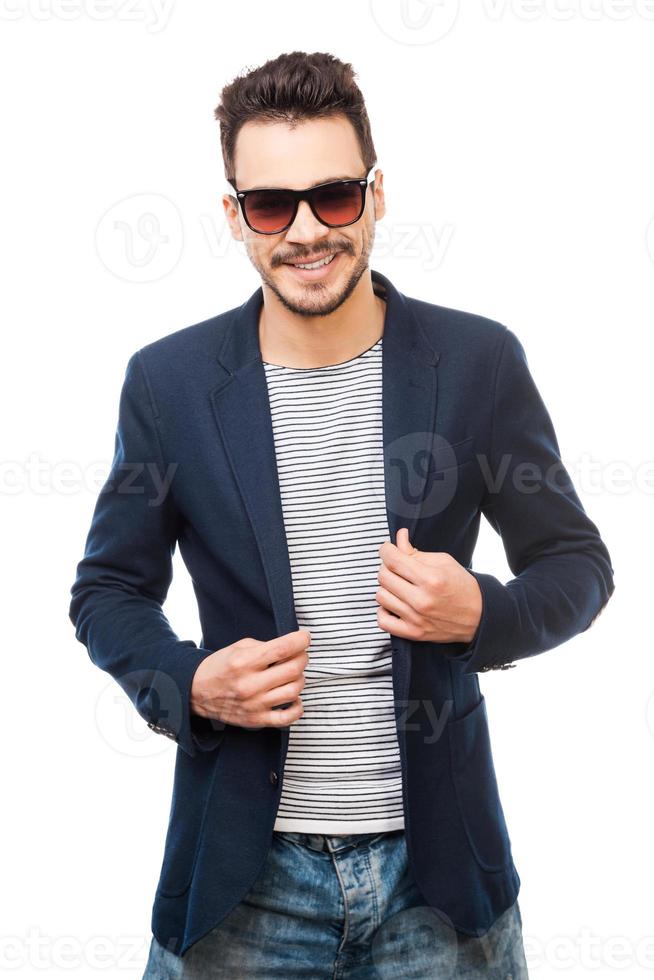 elegant und stilvoll. hübscher junger Mann mit Sonnenbrille, der seine Jacke anpasst, während er vor weißem Hintergrund steht foto