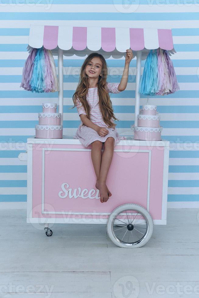 schönes Kind. süßes kleines Mädchen, das in die Kamera schaut und lächelt, während es auf der Dekoration des Süßigkeitenwagens sitzt foto