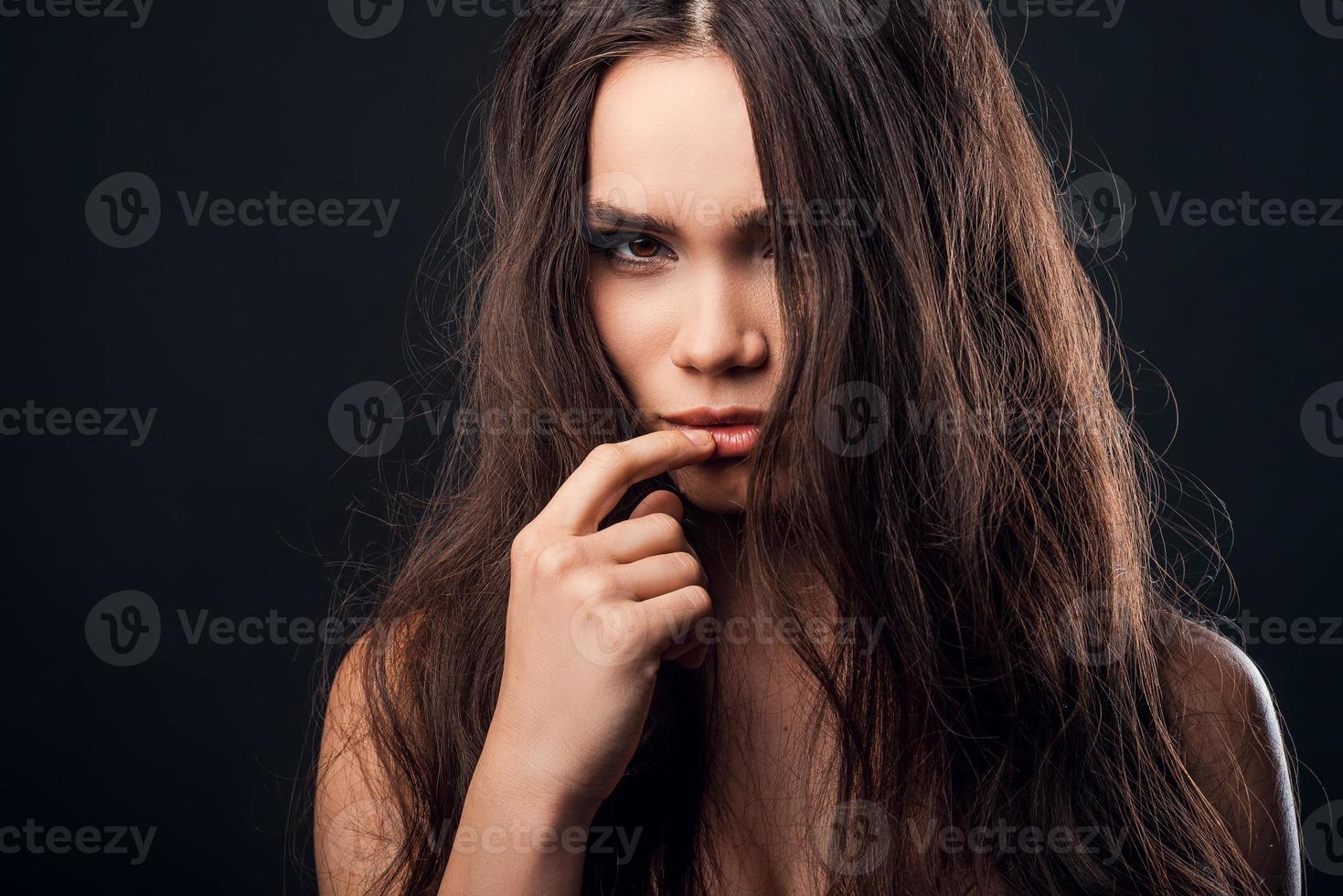 durchdringender Blick. selbstbewusste junge hemdlose Frau, die den Finger auf ihre Lippen hält und in die Kamera schaut, während sie vor schwarzem Hintergrund steht foto