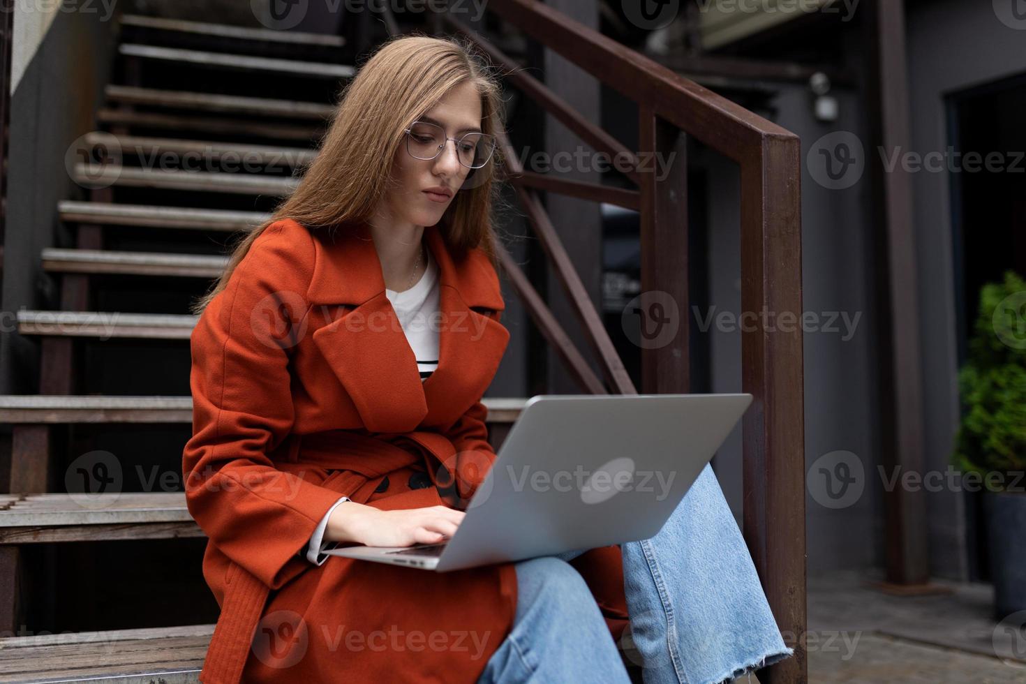 Porträt einer starken Studentin im Mantel, die mit einem Laptop auf dem Schoß auf der Treppe sitzt foto